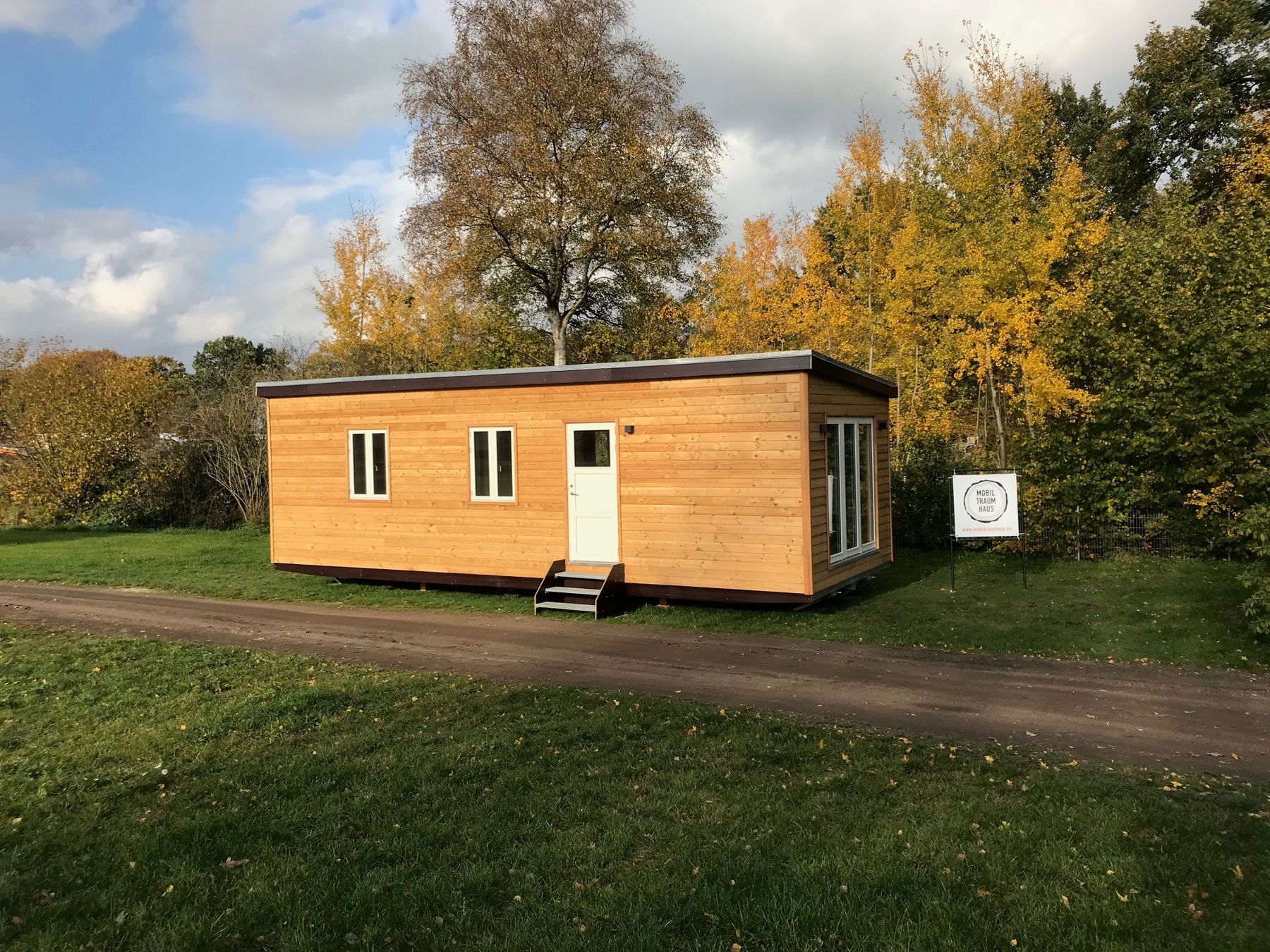 Ein Tiny House aus Holz auf einer Wiese vor herbstlichen Bäumen